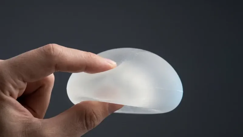 A close-up of a hand gently squeezing a translucent Gummy Bear Silicone Implant against a dark grey background to demonstrate its firm, cohesive texture.