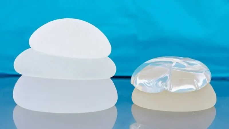 A side-by-side visual comparison of saline vs silicone breast implants, showing a stack of three opaque white implants on the left next to a clear, transparent implant resting on an opaque base on the right, all set against a blue background.